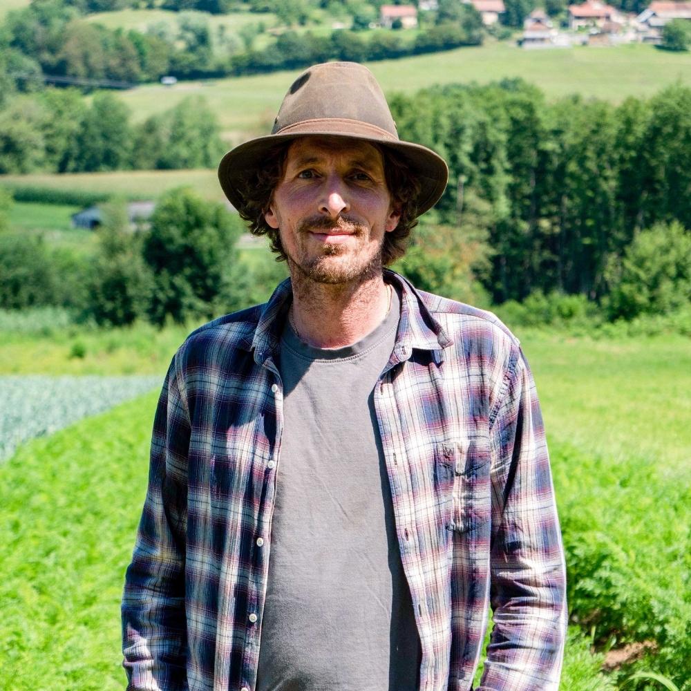 Portrait d'un agriculteur Trikome portant un chapeau, posant devant les cultures de chanvre bio avec un village de montagne en arrière-plan.